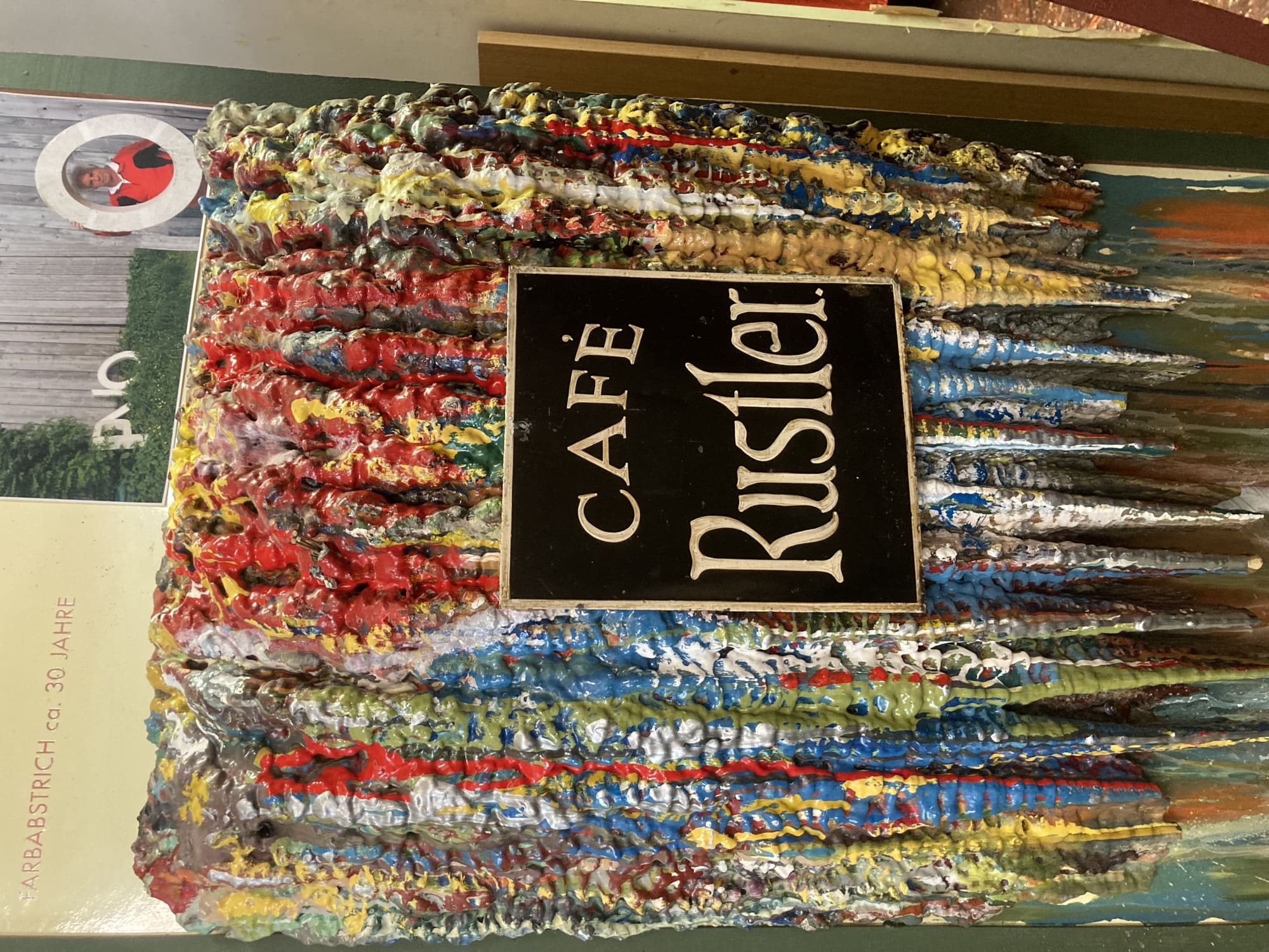 ein Objekt aus vielfarbigen Teilen, die nach unten wegzurinnen scheinen, in der Mitte ein schwarzes Schild mit der goldenen Aufschrift ”CAFÉ Rustler“