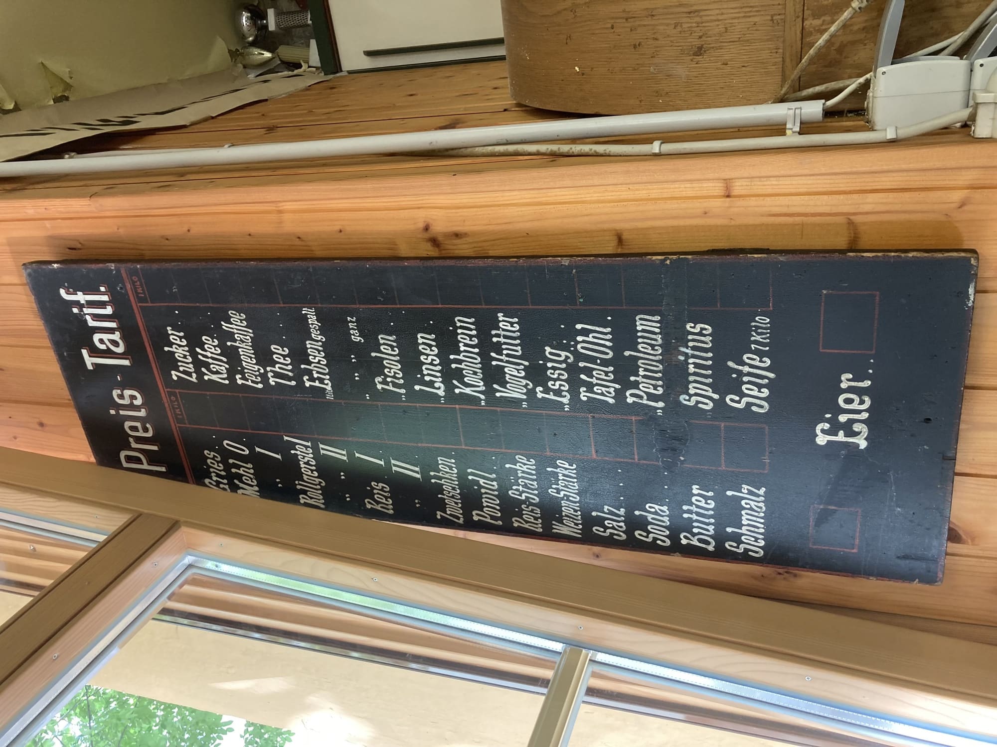 neben einem Fenster hängt ein schwarzes Schild mit der Überschrift „Preis-Tarif“, darunter sind diverse Produkte aufgelistet, daneben immer ein freier Platz für den Preis, Produkte sind zum Beispiel Mehl, Feigenkaffee, Butter, Thee (!), Tafel-Öhl (!) oder Petroleum