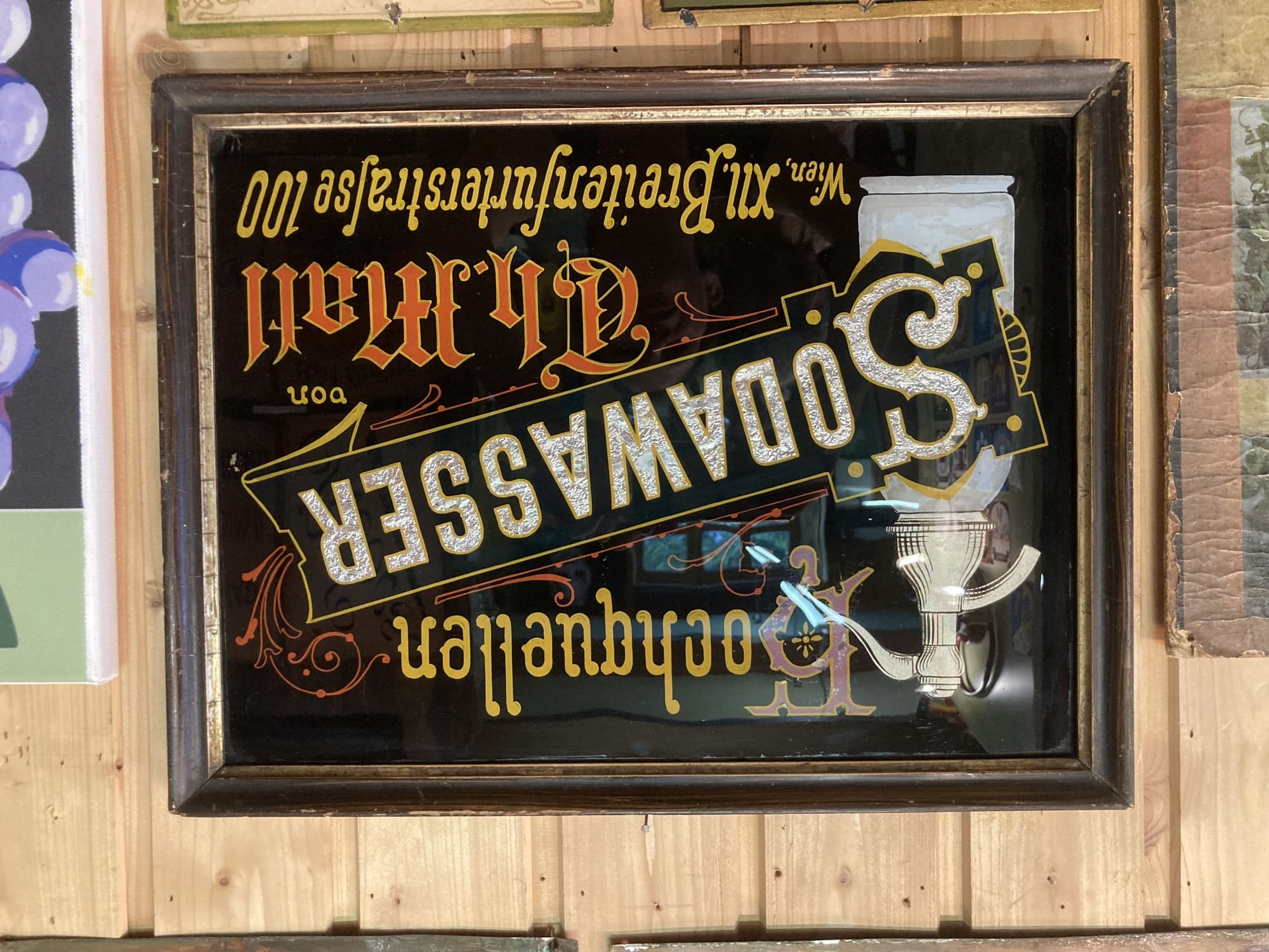 ein gemaltes Schild mit der Aufschrift „Hochquellen Sodawasser“, der Text Sodawasser ist schräg geschrieben und glänzt silbern auf schwarzem Hintergrund