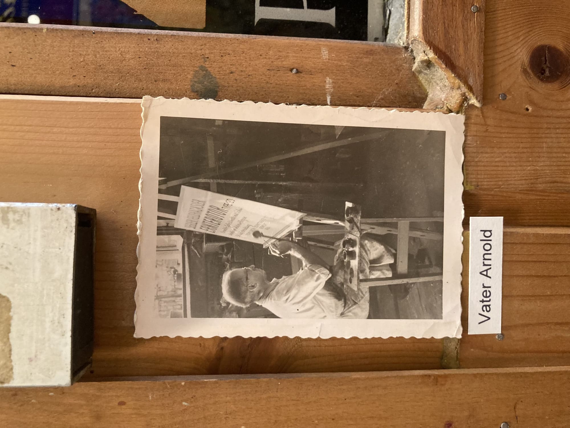 ein schwarz-weiß-Foto mit ausgefranstem Rand, das an einer Holzwand befestigt ist, auf dem Foto ist ein Mann zu sehen, der an einer Staffelei sitzt und an einem Schild malt, darunter der Text „Vater Arnold“