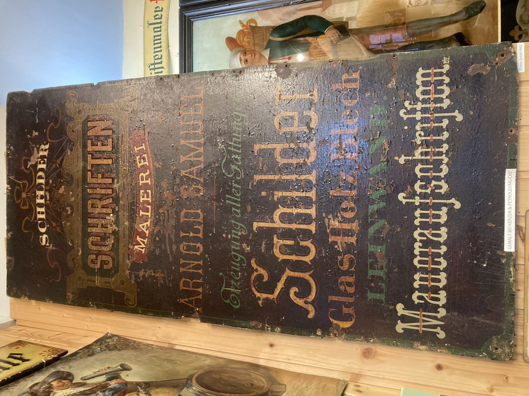 ein hohes Schild mit Texten in vielen unterschiedlichen Schriften, Texte von oben nach unten: „Schilder, Schriften, Malerei, Arnold Samuel, Erzeugung aller Gattungen, Schilder, Glas Holz Blech, Leinwand, Maueraufschriften“, darunter klein „gemalt bei Kerzen & Petroleumlicht“