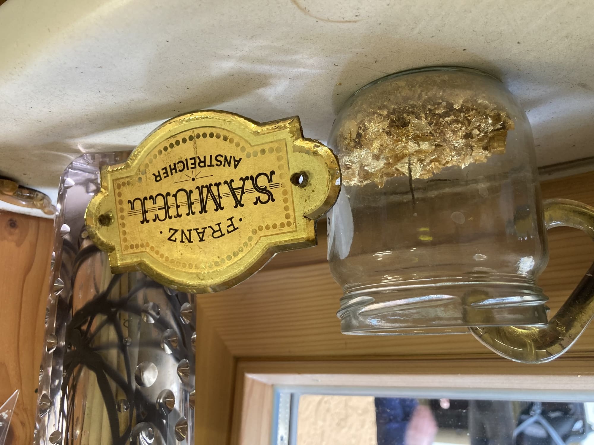 Nahaufnahme eines Glases mit Blattgoldfragmenten, daneben ein goldenes Schild mit dem Text „Franz Samuel Anstreicher“, der Text ist schwarz in drei verschiedenen Schriften auf goldenem Hintergrund, rundherum verziert mit einer Bordüre aus goldenen Punkten