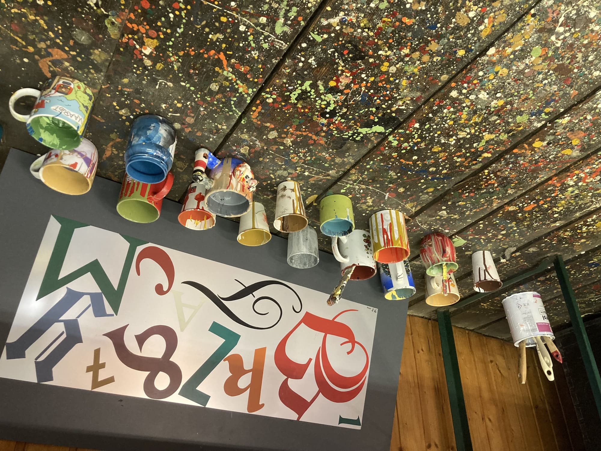 Musterblatt mit verschiedenen Buchstaben in verschiedenen Schriften und Farben, davor eine Sammlung von Bechern und Tassen, in denen verschiedene Farben angerührt wurden, im Vordergrund der Holzboden des Raums, darauf viele Farbspritzer in den unterschiedlichsten Farben