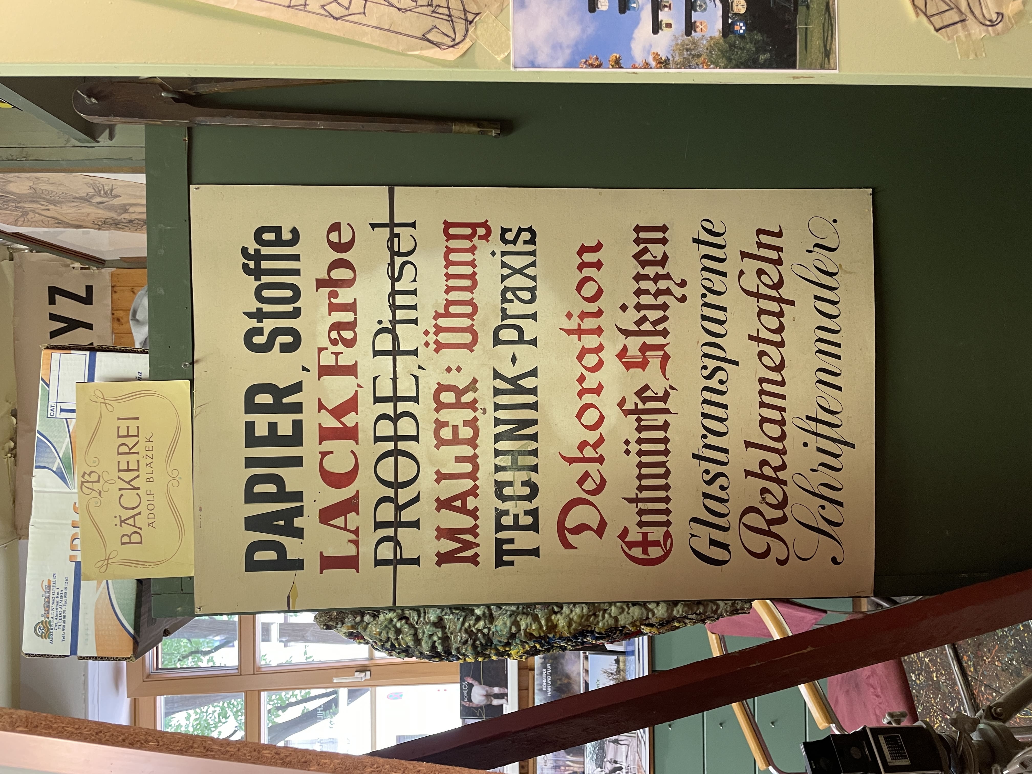 Tafel, die der Kundschaft des Schildermalers die verfügbaren Schriften zeigt, jedes Wort ist in einer anderen Schrift geschrieben, Farbe wechselt zwischen Schwarz und verschiedenen Rot-Tönen, Text: „Papier, Stoffe, Lack, Farbe, Probe, Pinsel, Maler: Übung, Technik-Praxis, Dekoration, Entwürfe, Skizzen, Glastransparente, Reklametafeln, Schriftenmaler.“