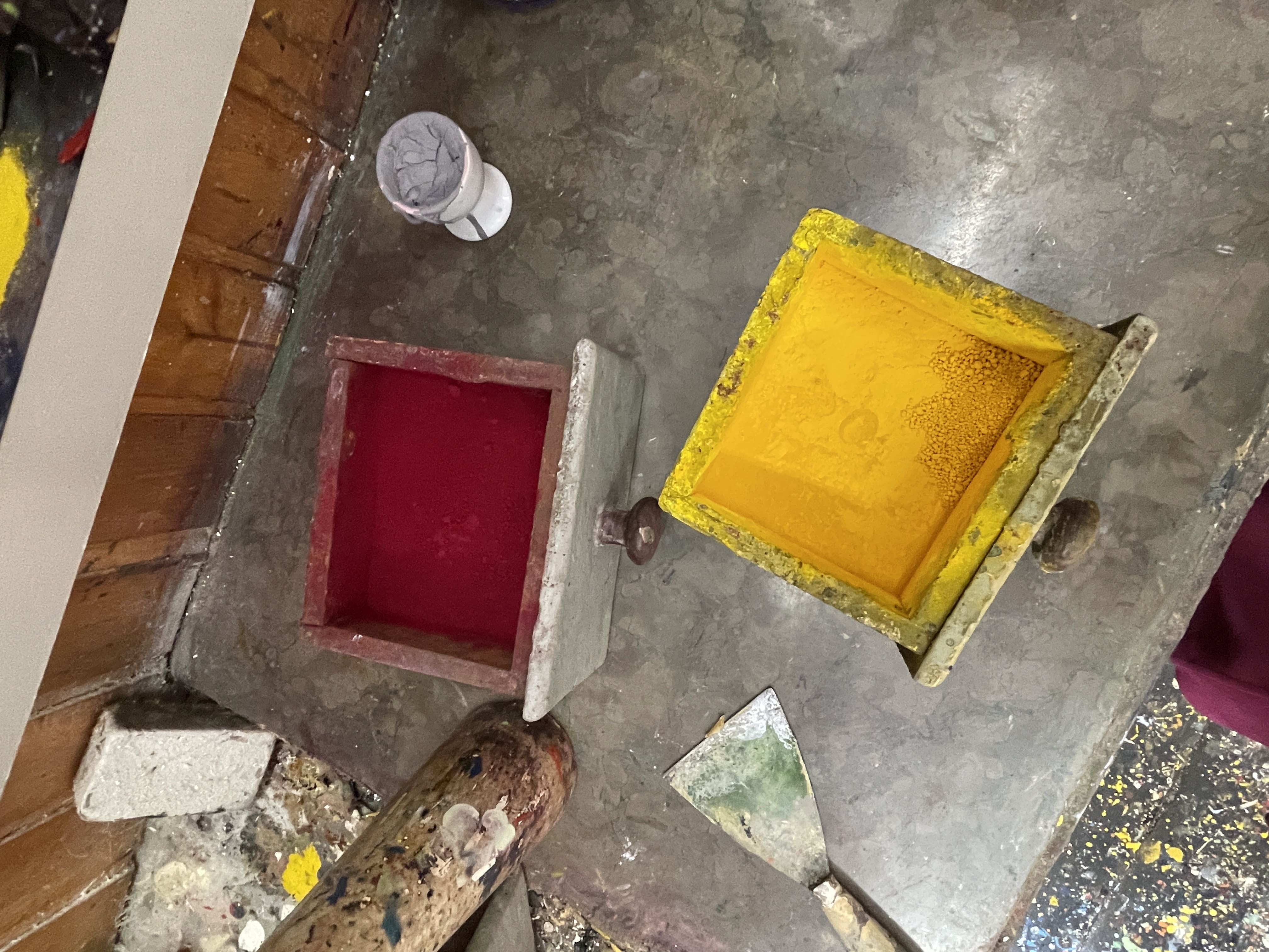 Blick von oben auf die Werkbank des Schildermalers, darauf stehen zwei offene Schubladen mit Farbpigmenten, oben bordeauxrot, darunter chromgelb, links daneben ein Farbspachtel, rechts oben ein Eierbecher mit einem grauen Belag