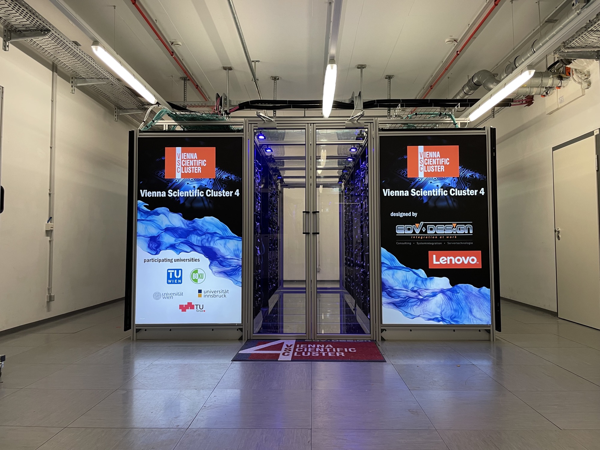 ein System aus Racks frei stehend in einem Raum, in der Mitte zwischen den Racks führt ein Gang hindurch, die Seitenflächen, die zur Kamera zeigen sind mit Vienna Scientific Cluster 4 und den Logos verschiedener beteiligter Firmen beschriftet