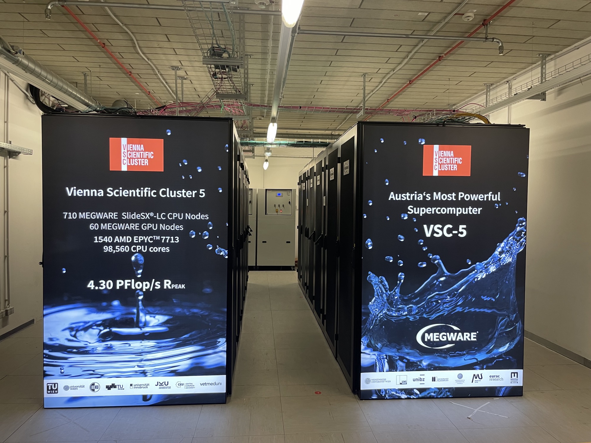 ein System aus Racks frei stehend in einem Raum, in der Mitte zwischen den Racks führt ein Gang hindurch, die Seitenflächen, die zur Kamera zeigen sind mit Vienna Scientific Cluster 5 und den Spezifikationen beschriftet