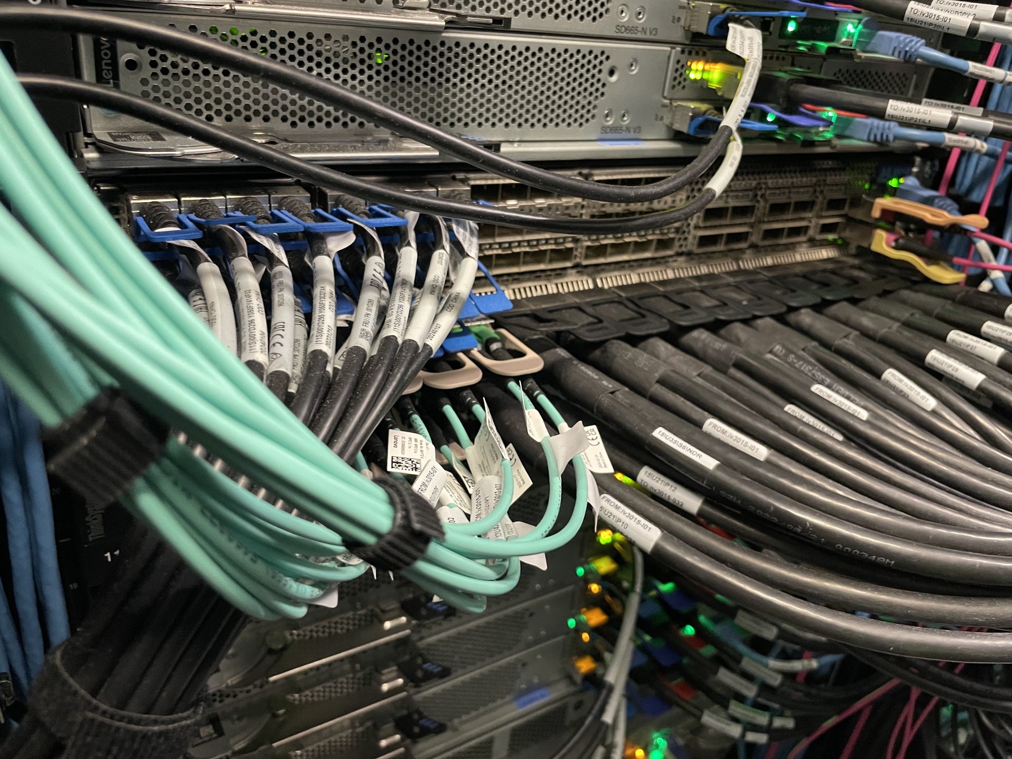 eine große Menge Kabel führt in ein Rack hinein, dünnere türkisfarbene Kabel (Lichtwellenleiter) in einem Bündel neben dickeren schwarzen Kabeln (Direct-attached-Copper (DAC), für kurze Netzwerk Strecken), rechts oben sind Netzwerkkabel zu erkennen