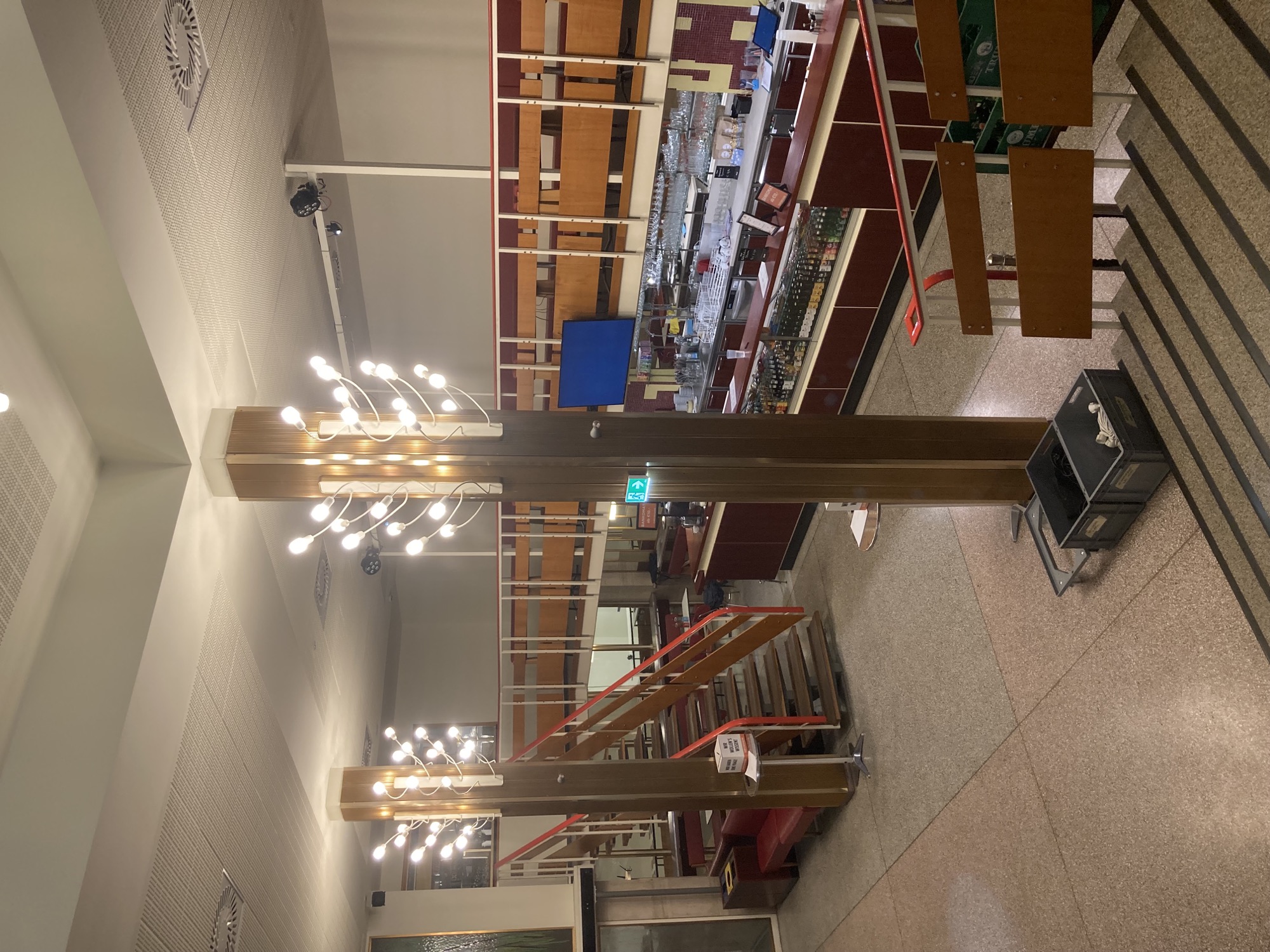 im Foyer des Kinos dominieren zwei hohe Säulen mit Lampenelementen, unter einer Galerie ist der Barbereich sichtbar, die dominierenden Farben sind Holz, Gold und verschiedene Rottöne